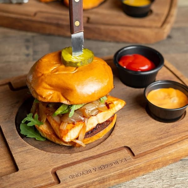 KX Burger + Beyond salmon burger with caramelized onions, arugula, and a pickle on a wooden board.