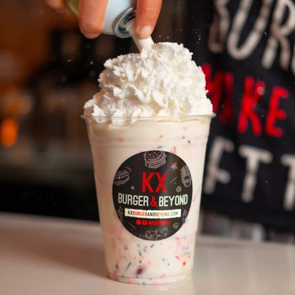 A hand adds whipped cream to a KX Burger + Beyond milkshake with colorful sprinkles.