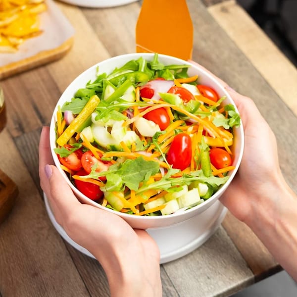 Hands holding a white bowl filled with a fresh salad of greens, tomatoes, cucumbers, and carrots.