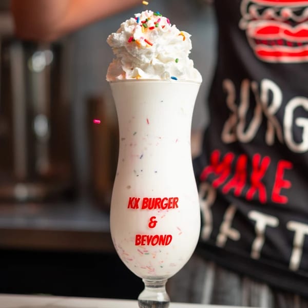 A KX Burger + Beyond milkshake with whipped cream and colorful sprinkles falling on top.