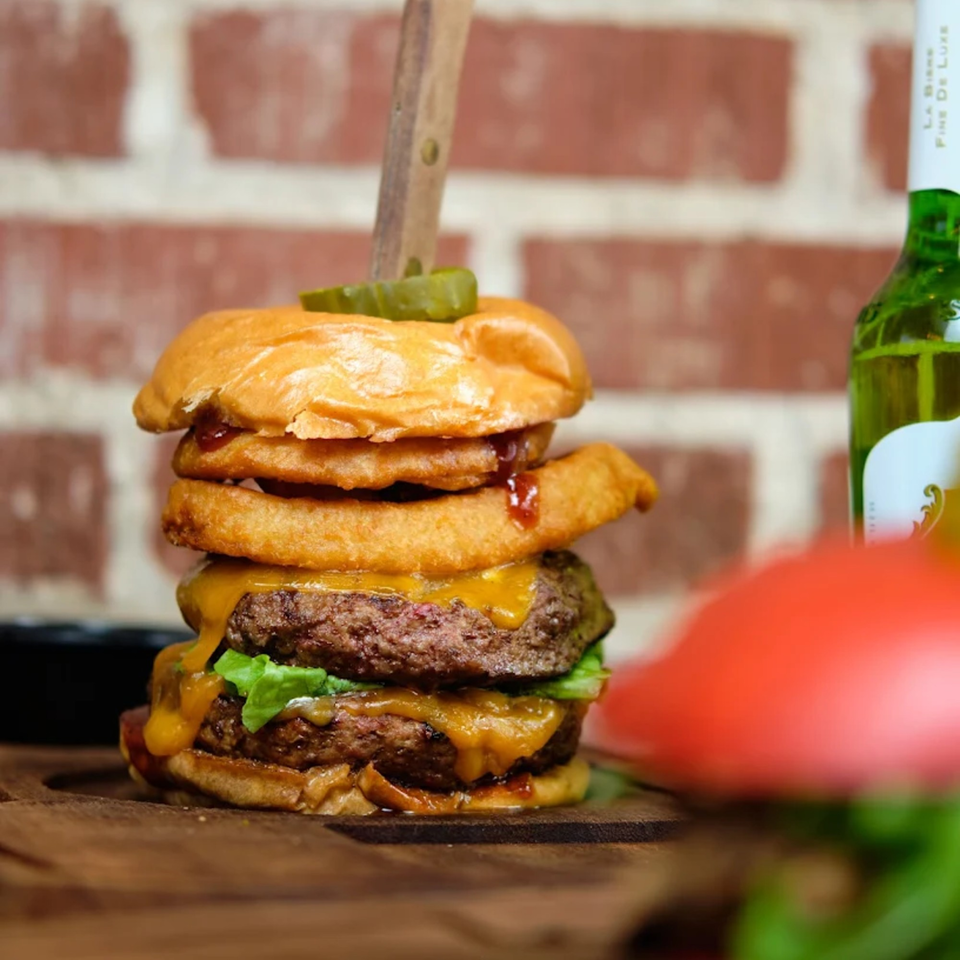 A double cheeseburger with onion rings, lettuce, and a pickle on a wooden board.