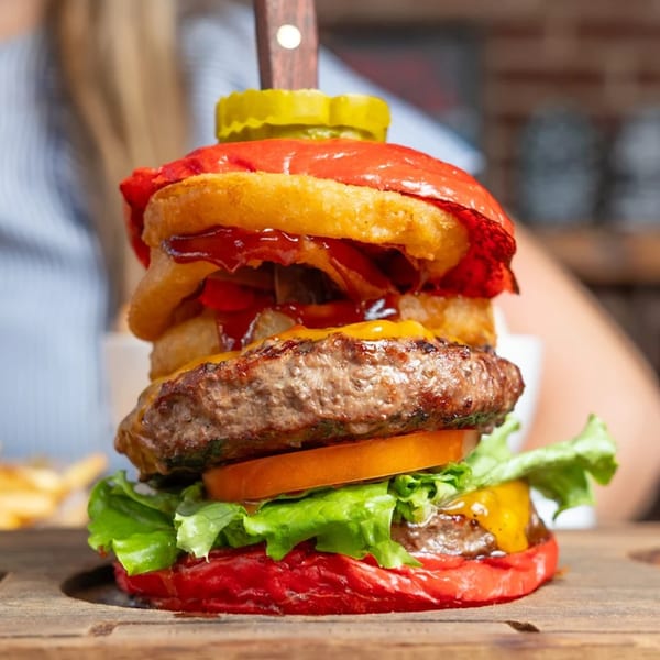 A KX Burger with a red bun, onion rings, bacon, cheese, lettuce, tomato, and a pickle.