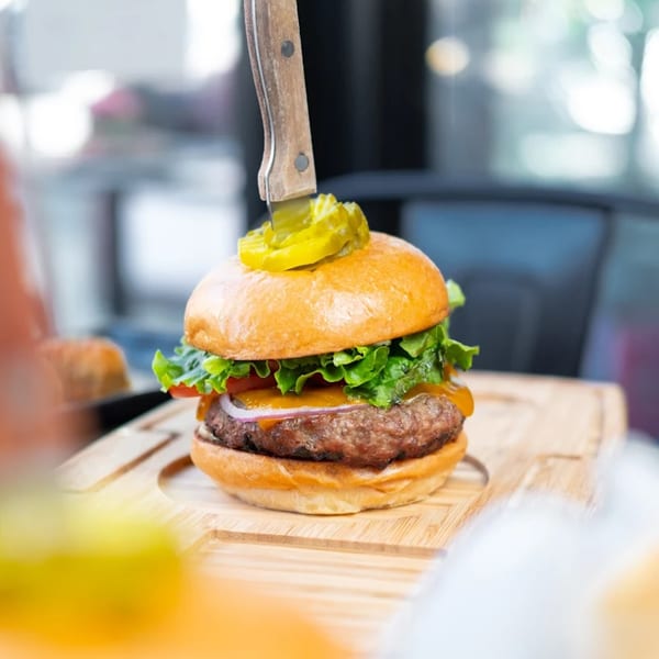 A KX Burger with cheese, lettuce, onion, and a pickle-topped knife on a wooden board.