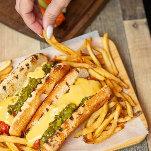 Two hot dogs with cheese and relish, and a side of fries, on a wooden board.